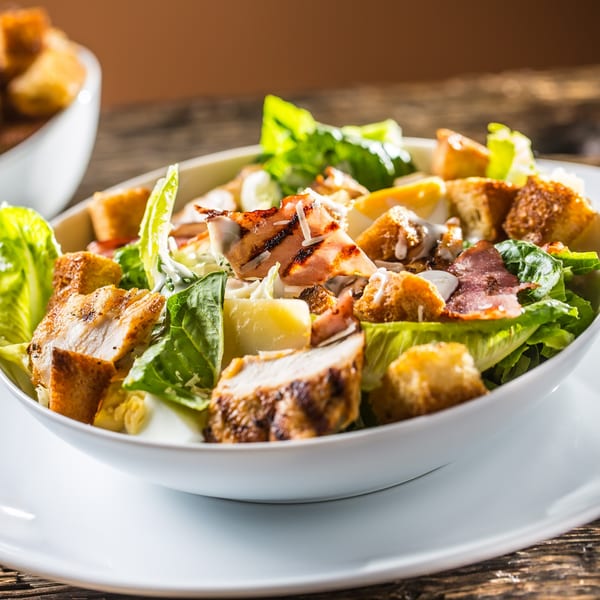 Grilled chicken Caesar salad with bacon, croutons, and dressing in a white bowl.