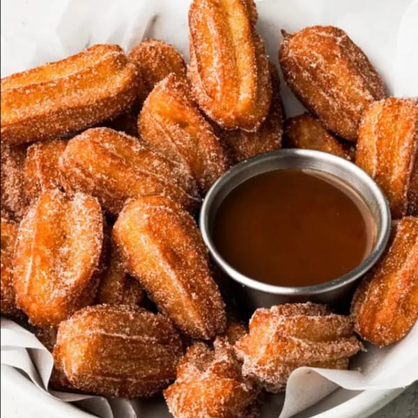 A white bowl filled with golden-brown churros dusted with sugar, served with a side of chocolate dipping sauce.