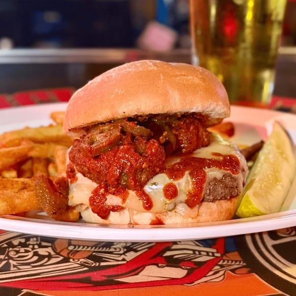A juicy burger with melted cheese, red sauce, and jalapeños, served with fries and a pickle.